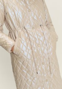 Cappotto beige lucido trapuntato con bottoni a pressione e lacci in vita, mani del modello nelle tasche laterali su sfondo semplice.