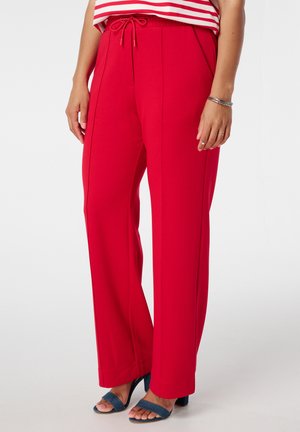 Rode, wijde broek van zachte stof met een tailleband met trekkoord. Bevat voornaad en wordt gedragen met blauwe haksandalen.