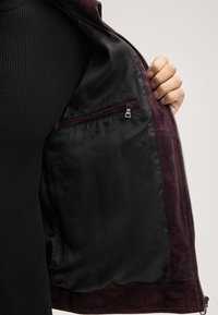 Interno di giacca scamosciata color vino con fodera in satin nero, dotata di tasca con zip e dettagli di cucitura visibili lungo il bordo.