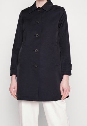 Personne portant un manteau noir boutonné à manches longues avec un col, associé à un pantalon blanc, debout devant un fond uni.