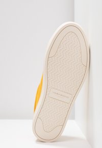 Semelle de chaussure jaune inclinée contre un coin blanc, bande de roulement beige à motif avec le logo « Vagabond Shoemakers » centré sur la semelle.