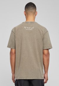Camiseta de algodón verde oliva con mangas cortas, que presenta texto impreso en blanco en árabe e inglés en la parte de atrás. Corte relajado, textura suave.