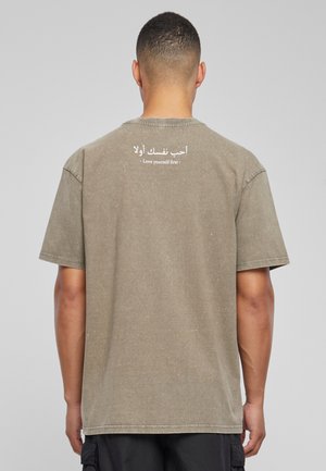 Olive green cotton t-shirt with short sleeves, featuring white printed text in Arabic and English on the back. Relaxed fit, soft texture.