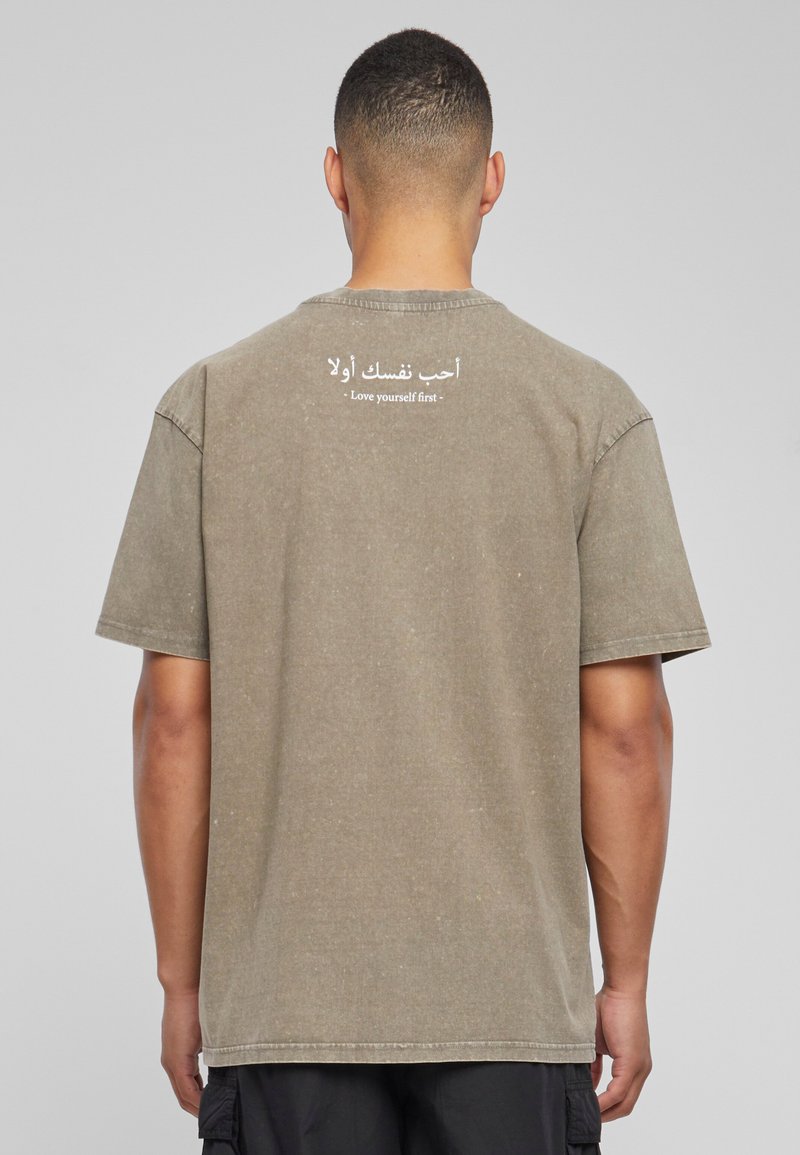 Camiseta de algodón verde oliva con mangas cortas, que presenta texto impreso en blanco en árabe e inglés en la parte de atrás. Corte relajado, textura suave.