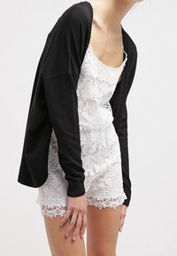 Svart kofta över en vit spetsromper. Koftan har långa ärmar och en avslappnad passform, medan rompen har delikat spets och vågiga kanter.