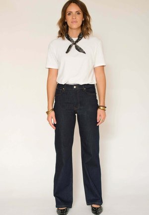 Jean taille haute bleu foncé avec une coupe droite, associé à un t-shirt blanc simple et une écharpe noire à motif. Chaussures à talons noires.