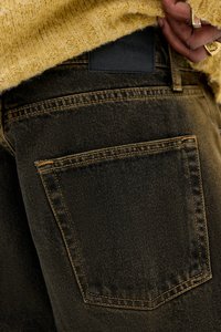 Nærbillede af en lomme på mørk denimbukser med synlig syning, båret med en gul tekstureret sweater, mens en hånd med guldringe justerer sweateren.