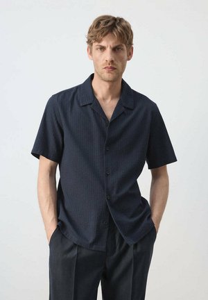 Jonge man met lichtbruin haar, gekleed in een donkerblauw kort mouw knoopshirt en zwarte broek met handen in de zakken.