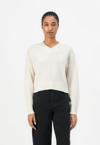 Pull court de couleur crème en tricot doux avec un décolleté en V et un ourlet côtelé, présentant un petit accent en forme de cœur sur la poitrine gauche.