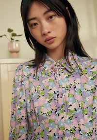 Vestido floral com um padrão multicolorido em rosa, verde e lavanda, sobre um fundo azul claro, com um colarinho estruturado e fecho de botões.