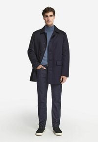 Marinfärgad kappa med knappar, bröstficka och skräddarsydd passform, kombinerad med en ljusblå polotröja, mörka denimbyxor och svarta sneakers.