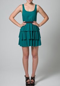 Türkisfarbenes ärmelloses Kleid mit tailliertem Oberteil, gestuftem Rüschenrock und einem schwarzen Taillengürtel. Kombiniert mit schwarzen offenen High Heels.