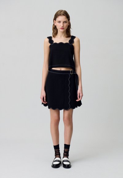 Crop top noir avec des bords festonnés, associé à une jupe noire avec un ourlet festonné et une ceinture latérale avec une boucle en or. Chaussures plates.