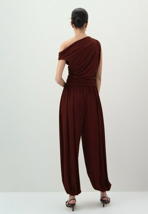 Woman with hair in a bun wearing a burgundy one-shoulder top and matching loose high-waisted pants with gathered hems, standing facing away.