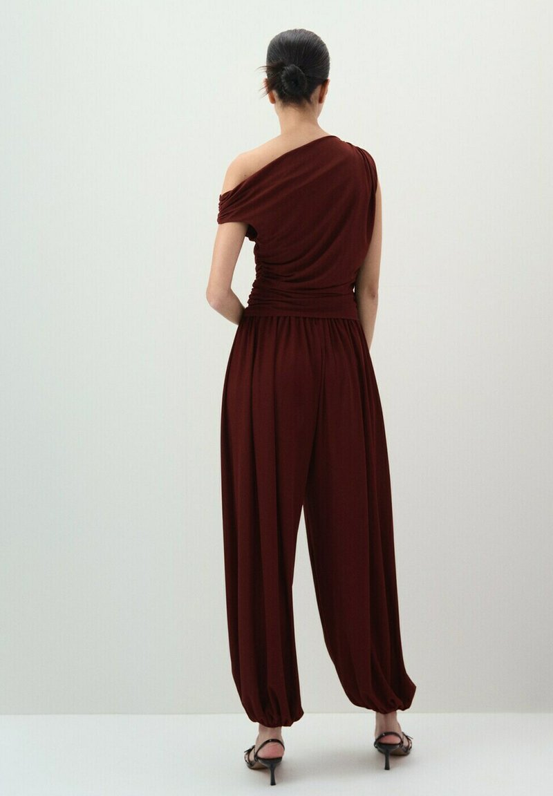 Femme avec les cheveux en chignon portant un haut bordeaux à une épaule et un pantalon ample taille haute assorti avec des ourlets froncés, debout de dos.