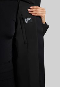 L'abbigliamento esterno nero presenta una texture morbida, tasche nascoste e un'etichetta logo, con un design slim che mette in risalto linee e cuciture morbide.