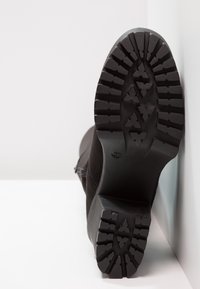 Bota de ante negra con una suela de goma texturizada, que presenta un patrón de banda de rodadura resistente y un ligero tacón, inclinada sobre una superficie blanca.