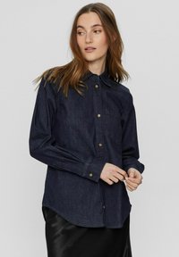 Mörk denimskjorta med krage, långa ärmar och en bröstficka. Har guldtonade knappar och en rundad nederkant.