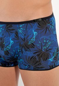Boxer-Briefs aus einem dehnbaren blauen Stoff mit einem schwarzen Blumenmuster und türkisfarbenen Akzenten sowie einem schwarzen Bund mit einem Etikett.