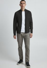 Giacca a zip grigia scura in maglia sopra una t-shirt bianca, abbinata a pantaloni beige chiari e sneakers nere. Design minimalista, texture evidenti.