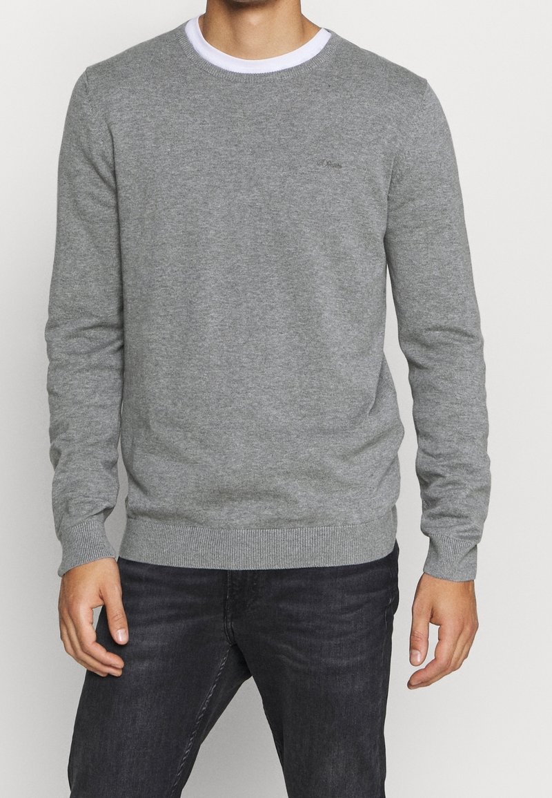 Grauer Pullover mit Rundhalsausschnitt, gerippten Bündchen und Saum. Aus weichem Material gefertigt. Auffälliges Logo-Detail auf der Brust. Kombiniert mit dunklen Jeans.