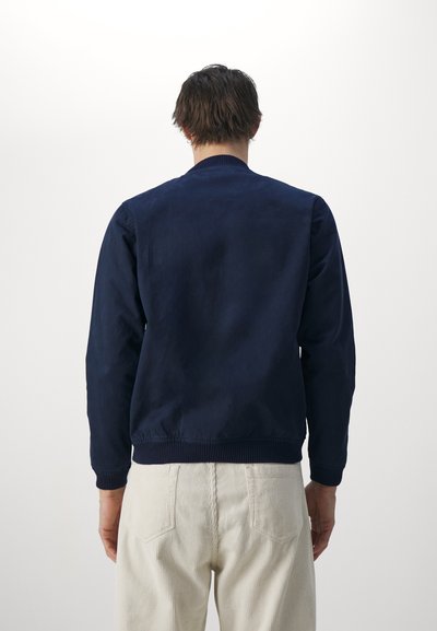 Chaqueta bomber azul marino de tejido suave, con cuello y puños de canalé. La parte trasera es sencilla, complementando unos pantalones de pana de color claro.