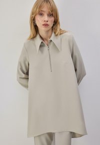 Tunique beige clair à fermeture éclair avec col pointu, manches longues et deux poches avant. Tissu lisse avec une coupe décontractée et ample.