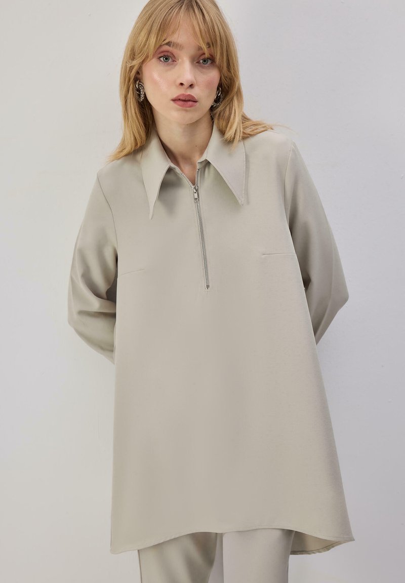 Tunique beige clair à fermeture éclair avec col pointu, manches longues et deux poches avant. Tissu lisse avec une coupe décontractée et ample.
