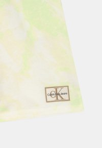 Hoek van een bleekgele en groene tie-dye stof met een beige en witte rechthoekige Calvin Klein Jeans-label gestikt nabij de rand.