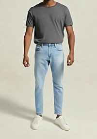 Homme portant un t-shirt à manches courtes gris foncé, un jean slim bleu clair et des baskets blanches, debout sur un fond uni.