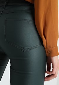 Tummanvihreät legginsit, joissa on sileä, kiiltävä pinta, takatasku ja kapea istuvuus, yhdistettynä pitkähihaiseen oranssiin paitaan.