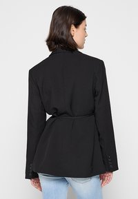 Blazer negro a medida con una cintura entallada, hombros estructurados, mangas largas y detalles de botones en los puños. Textura de tela suave.