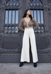 Cardigan à carreaux beige et marron avec des boutons, associé à un pantalon large crème. Bottines noires à talons. L'arrière-plan présente des portes en bois décoratives.