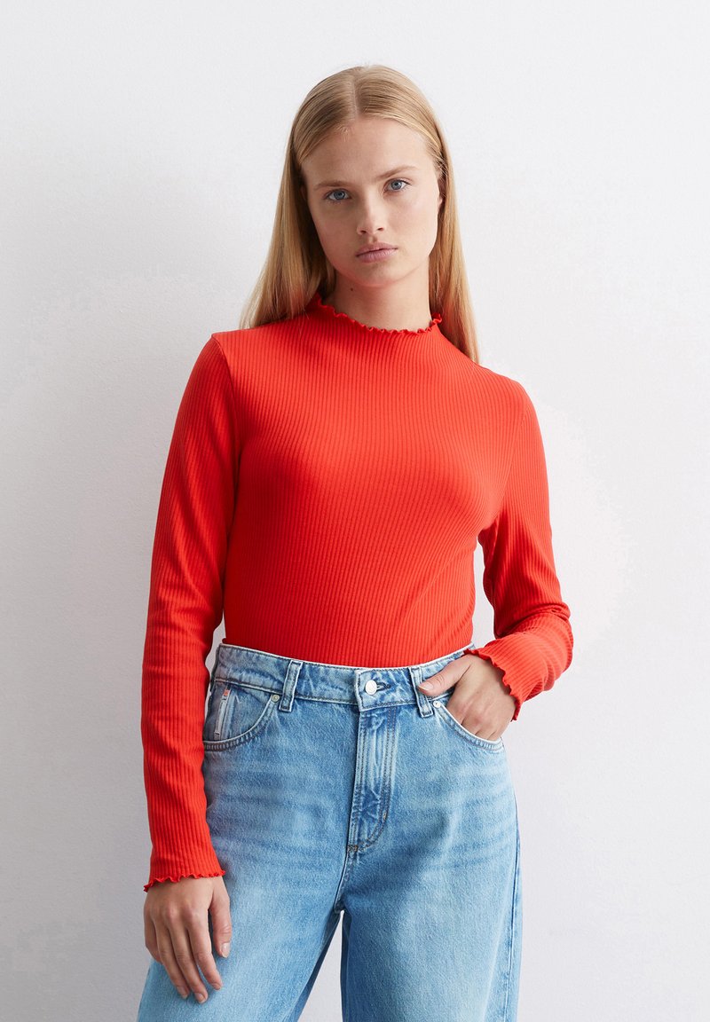Rotes, geripptes Langarmoberteil mit einem gefransten Halsausschnitt, kombiniert mit hoch taillierten, verblassten blauen Jeans. Glatte Textur und enganliegendes Design.