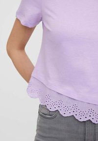 Fialový top s krátkým rukávem a vlnkovaným lemem, ozdobený vyšívanými detaily, v kombinaci se šedými denimovými džínami. Hladká textura a volný střih.