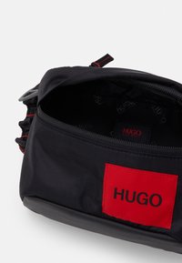 Sac textile noir avec fermeture éclair. Présente une patch logo rouge, des coutures contrastantes et une doublure intérieure avec des impressions de logo répétées.
