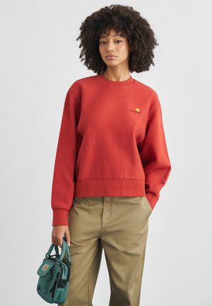 Γυναίκα με σγουρά μαλλιά που φοράει κόκκινο φούτερ και χακί παντελόνι, κρατώντας μια μικρή τσάντα σε τυρκουάζ χρώμα, στέκεται μπροστά σε απλό φόντο.