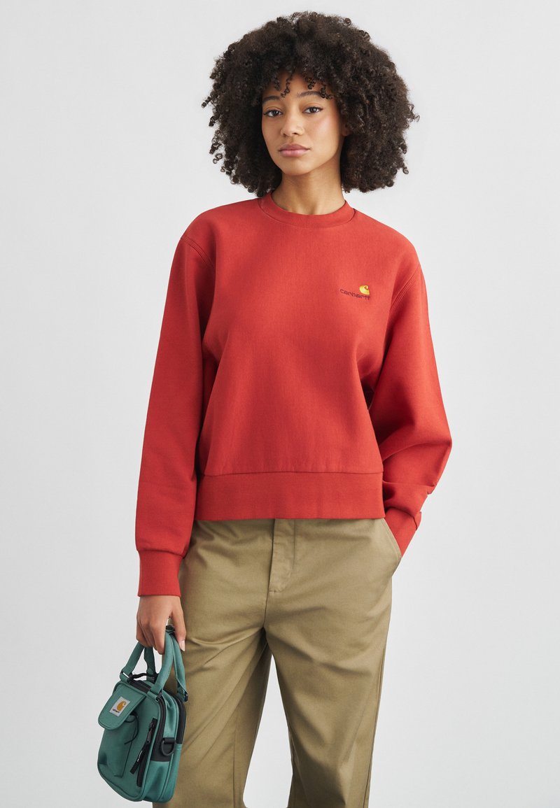 Γυναίκα με σγουρά μαλλιά που φοράει κόκκινο φούτερ και χακί παντελόνι, κρατώντας μια μικρή τσάντα σε τυρκουάζ χρώμα, στέκεται μπροστά σε απλό φόντο.