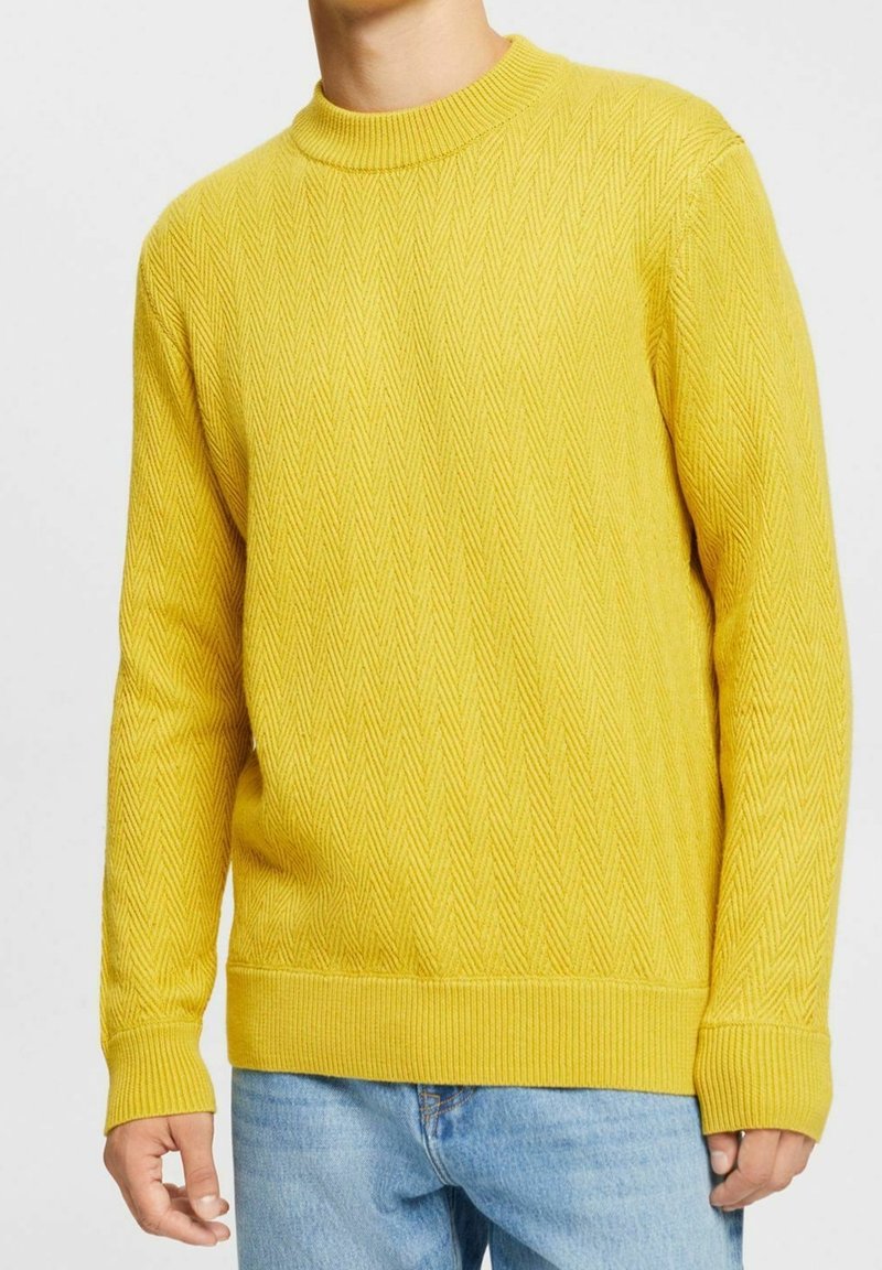 Homme portant un pull jaune texturé en tricot et un jean bleu clair, visible du cou jusqu'aux hanches sur un fond blanc.
