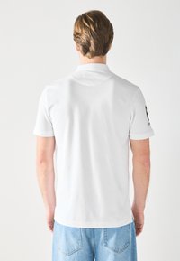 Polo blanc à manches courtes, texture lisse, avec un petit logo sur la manche gauche ; associé à un jean en denim bleu clair.