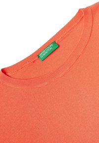 Korálové triko s krátkým rukávem vyrobené z měkké bavlny, se žebrovaným výstřihem a zelenou značkovou visačkou poblíž límce.