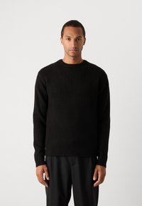 Pulover negru din tricot cu un guler rotund și mâneci lungi, realizat dintr-un material texturat. Purtați peste pantaloni negri, pe un fundal alb.