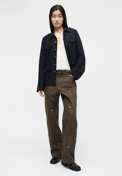 Γυναίκα που φοράει ναυτικό μπουφάν με κουμπιά, λευκό τοπ με άνοιγμα στο ντεκολτέ, καφέ παντελόνι με ασημί καρφιά, μαύρα παπούτσια και ζώνη με καρφιά.