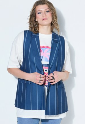 Donna con capelli mossi fino alle spalle indossa grandi orecchini a cerchio, un gilet blu a righe, una T-shirt bianca grafica, diversi anelli e jeans azzurri chiari.