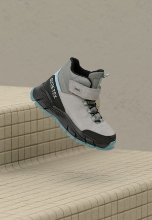 Bota de senderismo gris claro y negra con detalles azules, etiqueta Gore-Tex, correa de Velcro y suela texturizada, suspendida sobre escalones de baldosas.