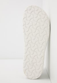 Semelle de chaussure blanche avec un motif texturé et ondulé ainsi qu'un logo de marque, affichant une forme arrondie et une finition lisse.