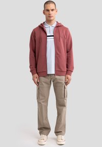 Bordeaux-roter Reißverschluss-Hoodie mit Fronttaschen, hellblaues Polohemd mit Streifen, beige Cargohose und weiße Schuhe mit roten Akzenten. Glatte Textur.