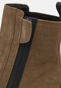 Brown suede ankle boot featuring a black zipper along the side, with raw edges and textured surface for added detail.