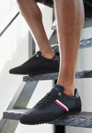 Persoon met zwarte sneakers met rood, wit en blauw gestreepte details, die een metalen diamantprofieltrap betreedt.