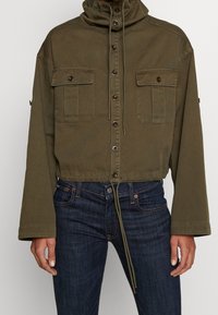 Avskuren, olivgrön jacka i bomullsblandning med hög krage, framknäppning och två bröstfickor. Dragsko nederkant och vida ärmar.